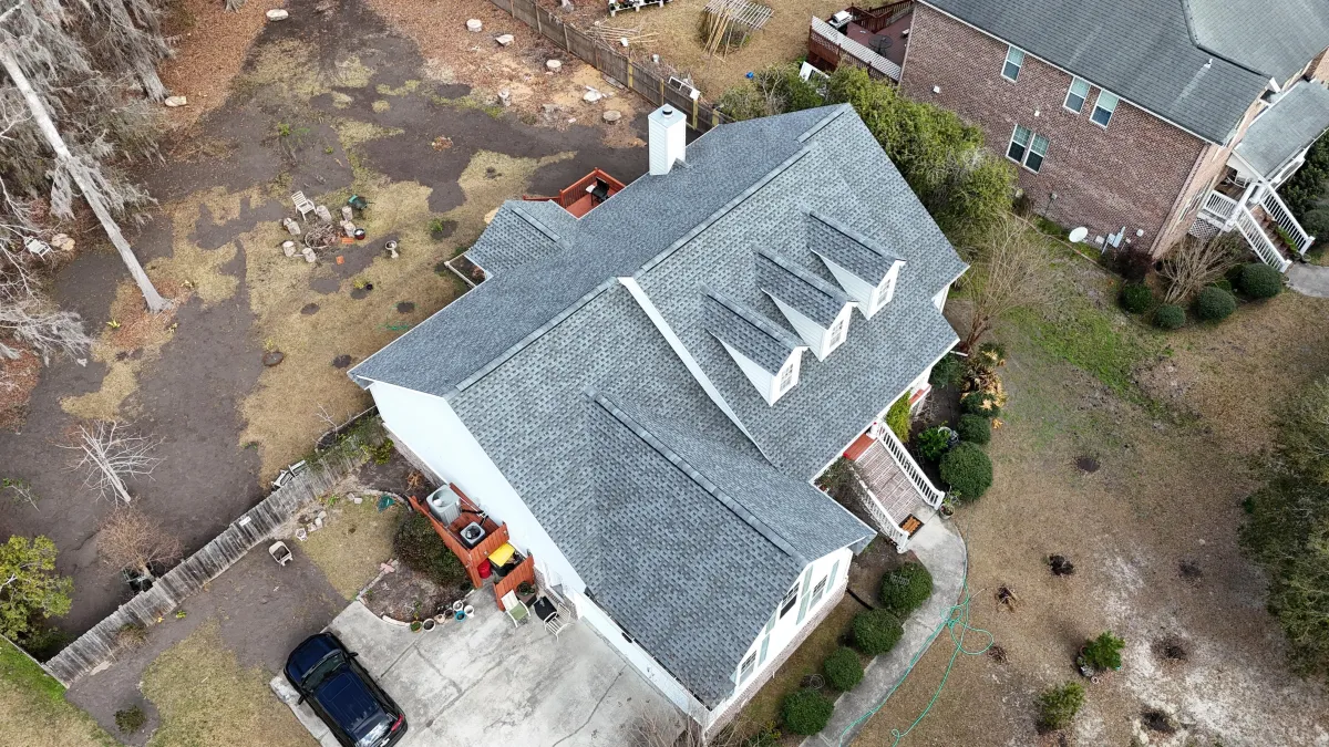 Aerial view of a residential property featuring a newly installed gray shingle roof, surrounded by landscaped yard with garden beds and a driveway, showcasing Precision Roofing USA's quality craftsmanship and attention to detail in roofing projects.