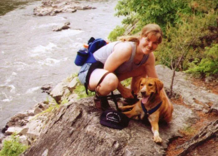 Young woman crouching next to her dog. Both are on a boulder next to running water..