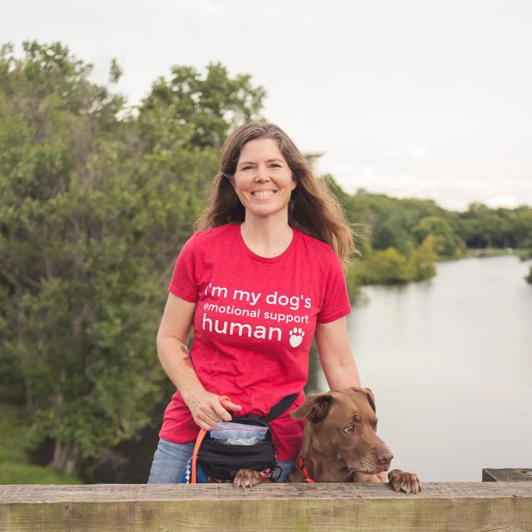 Woman kneeling next to her dog. Both are on a grassy area next to running water.