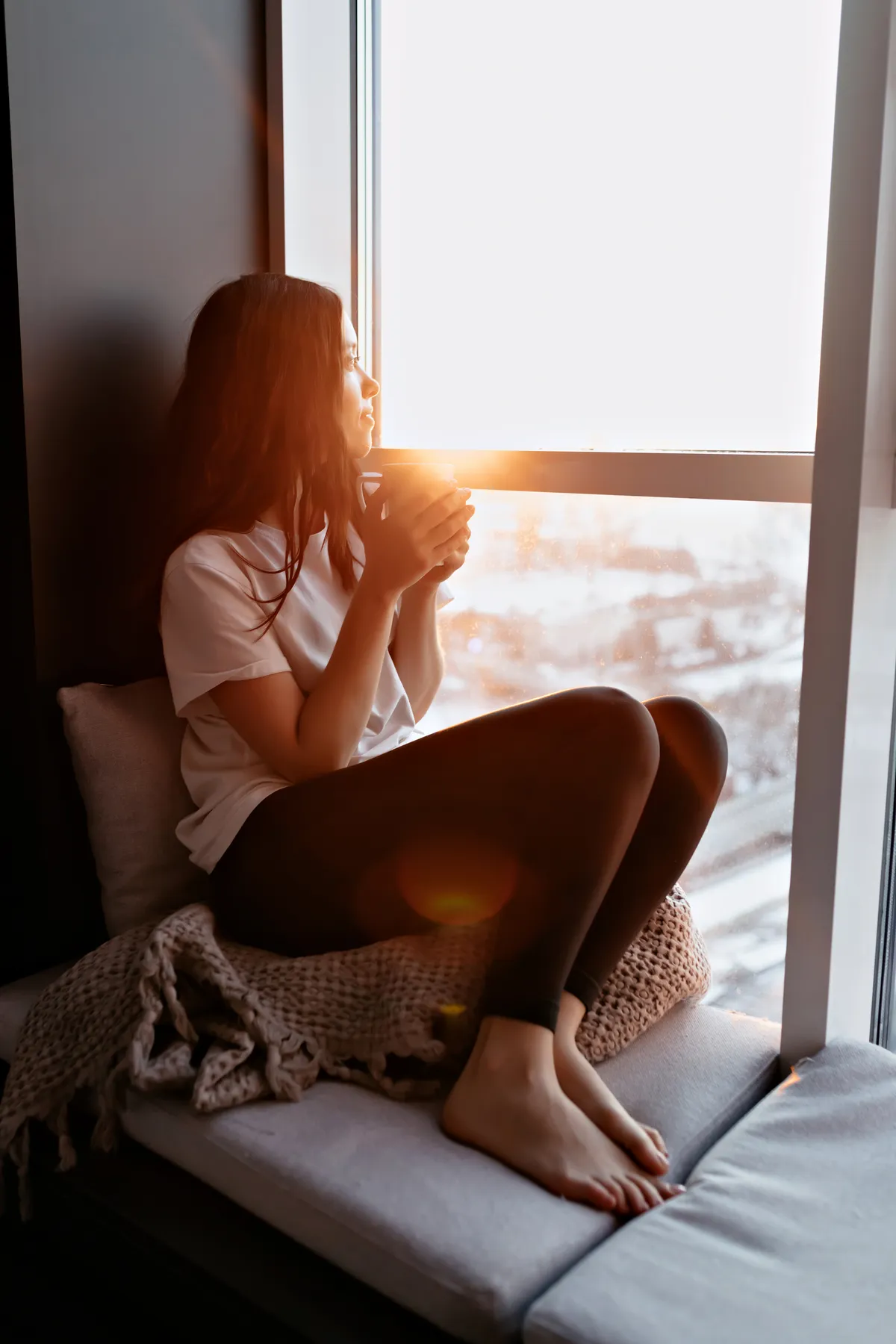 Woman sitting by a window holding a warm drink and looking outside