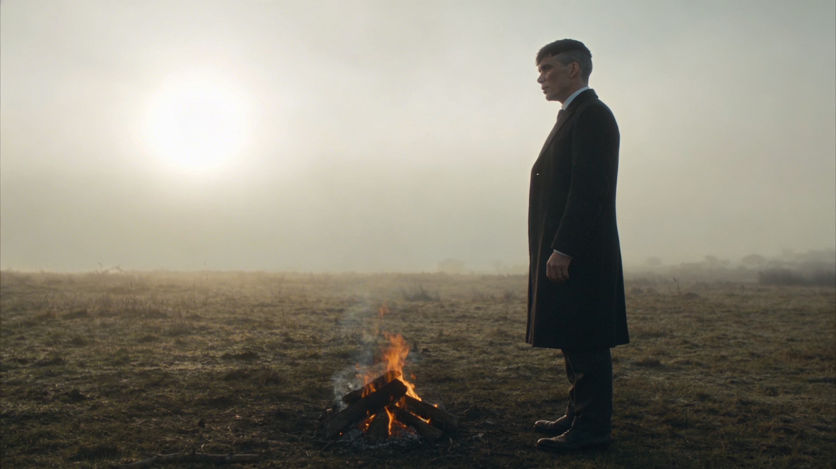 Solitary man in a dark coat standing beside fading embers in a misty landscape, reflecting a quiet release from control, survival and the life he once had to fight for.