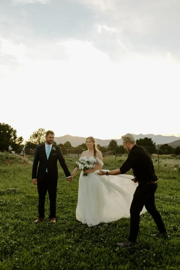 Bride and groom hold hands at sunset while a photographer adjusts the bride’s dress, mountains in the background. Wedding Videographer Arizona