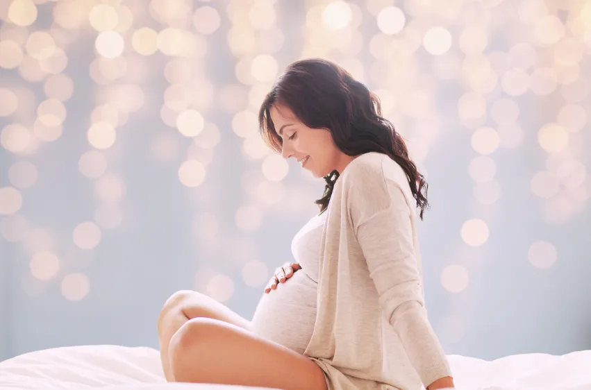 Pregnant woman sat on a bed cradling her baby bump. Calm, relaxed lighting in the background.