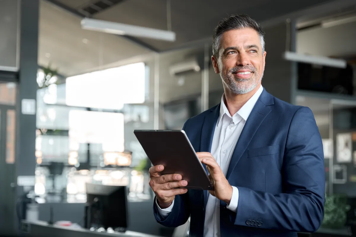 Middle-aged CEO in suit using a digital tablet in office, smiling and thinking while working on a tech device