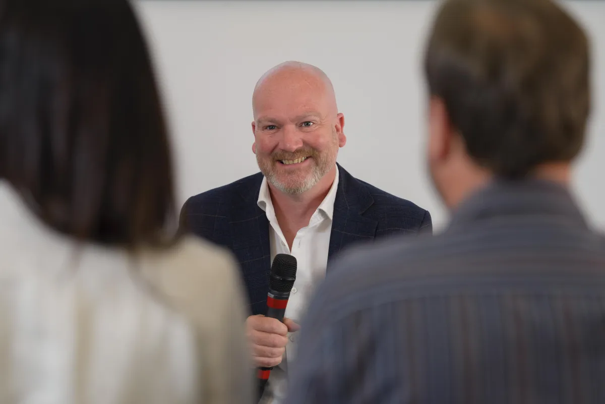 "Oliver Johnson, a professional business coach, smiling and holding a microphone while speaking to two attendees during a coaching session."