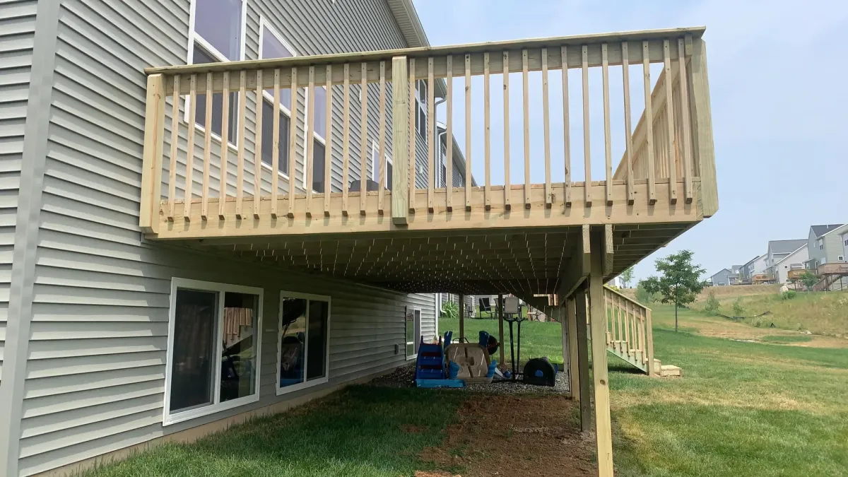 A spacious wooden deck elevated on posts, providing shaded space underneath, with a beige siding house in the background.