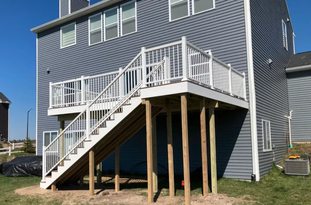 An elevated deck with white railings and a staircase, supported by wooden posts, attached to a gray siding house.