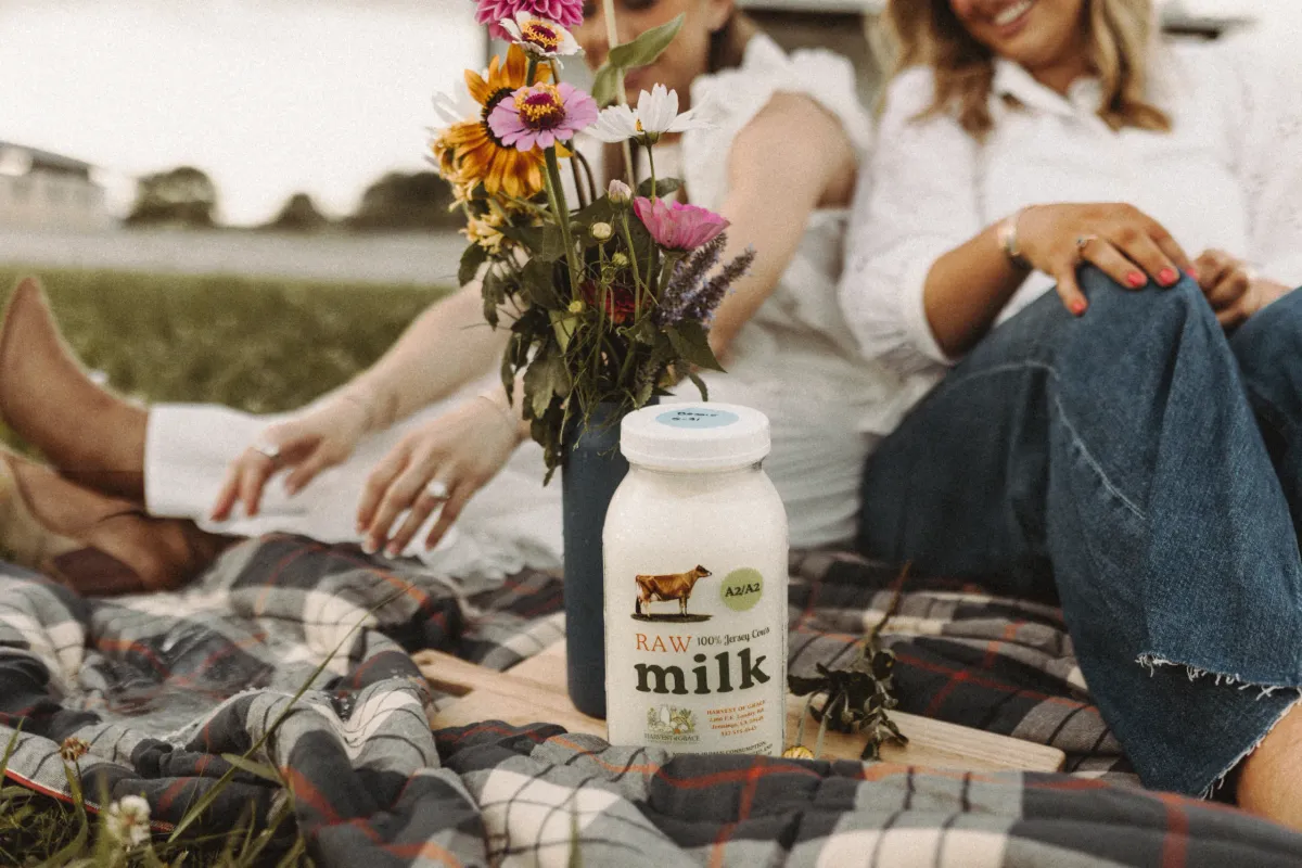 Two women share a drink on a blanket beside a grassy field.