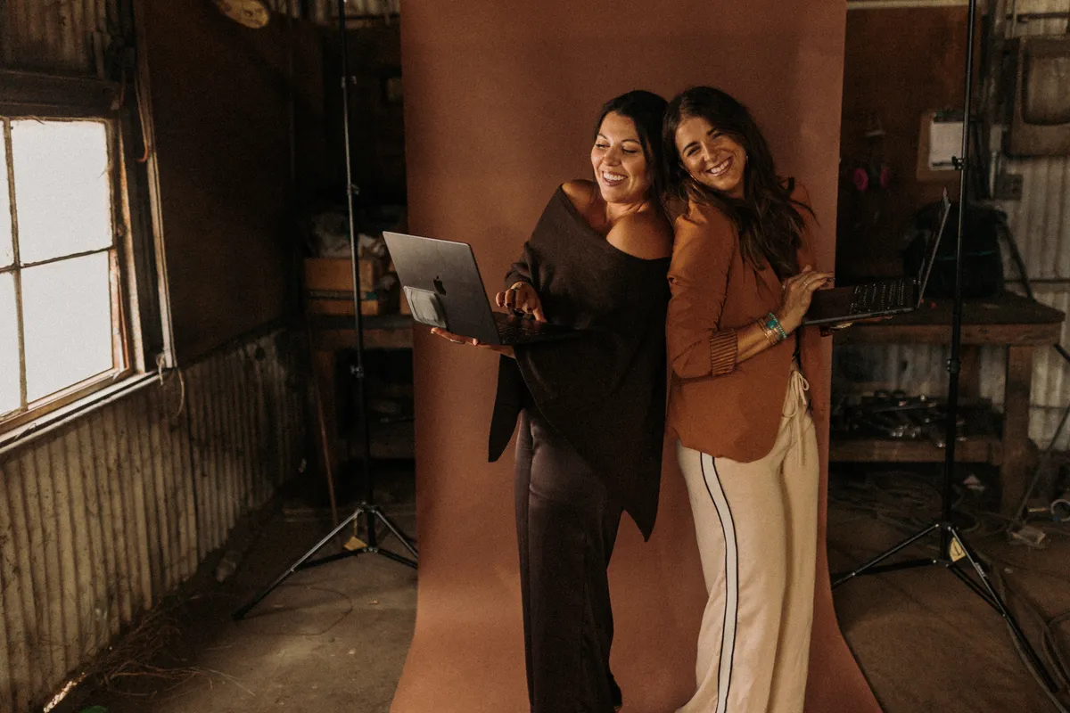 Courtney and Kori of Coyote Brands smiling with laptops in front of a studio backdrop