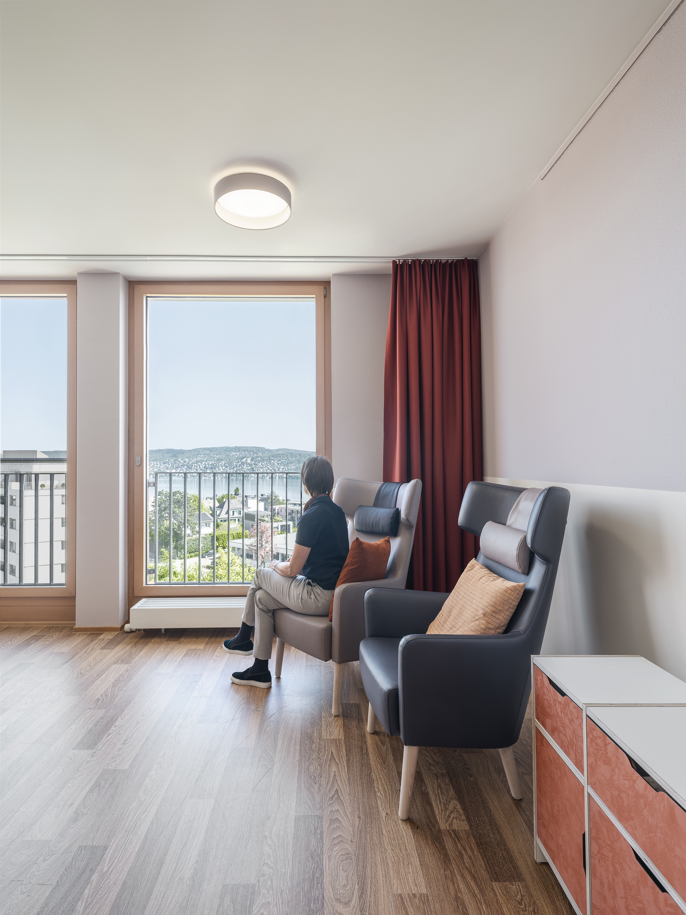 Wohnbereich für Menschen mit Demenz. Aufenthalt. Bewohnerin sitzt auf Ohrensessel. Blick aus dem Fenster auf Zürichsee