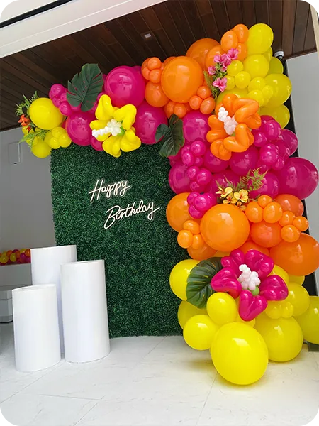 Tropical-style balloon garland with yellow and green tones over greenery wall