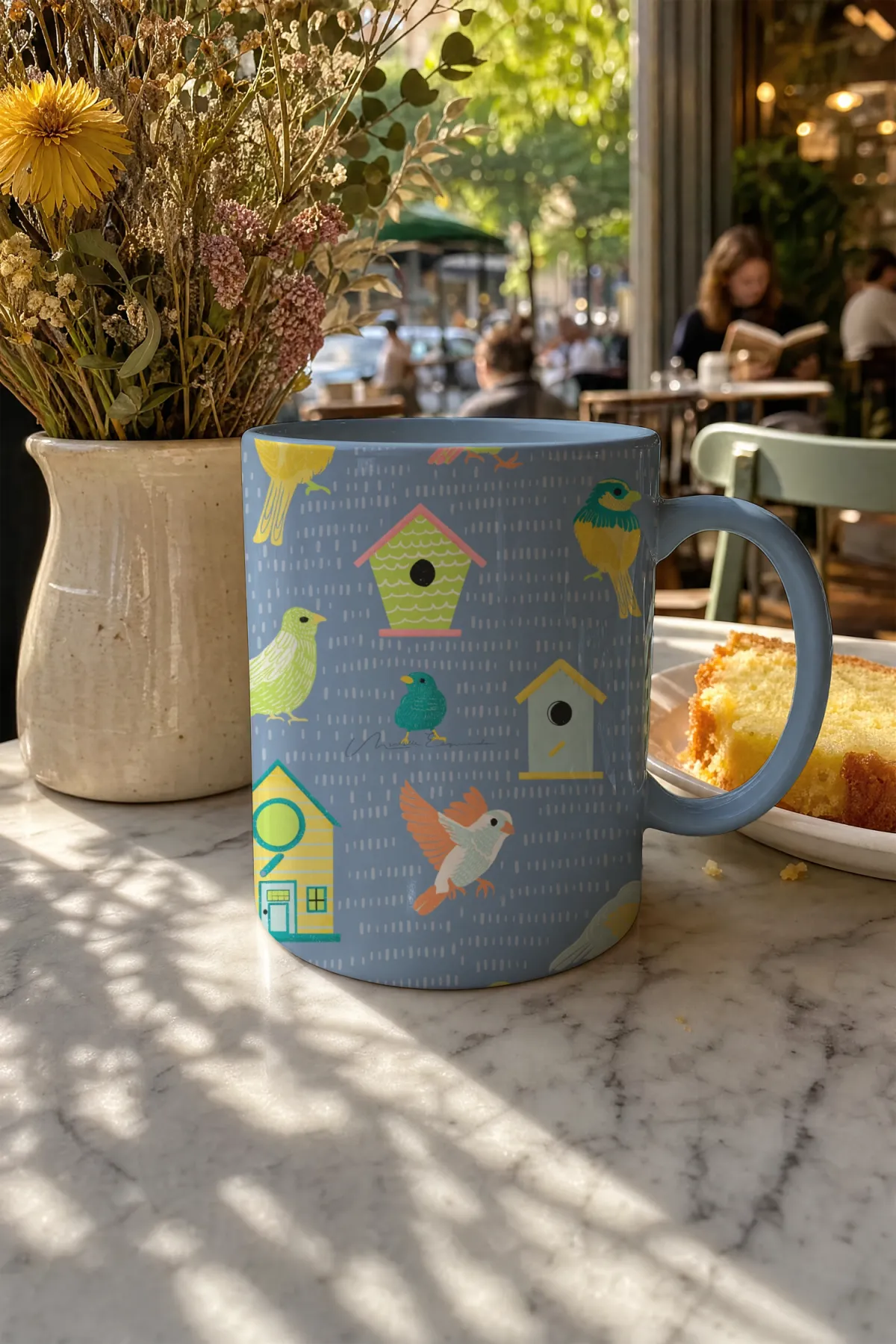 Coffee mug with the Home Tweet Home bird repeat pattern, styled on a marble table in a restaurant with a plate of cake.