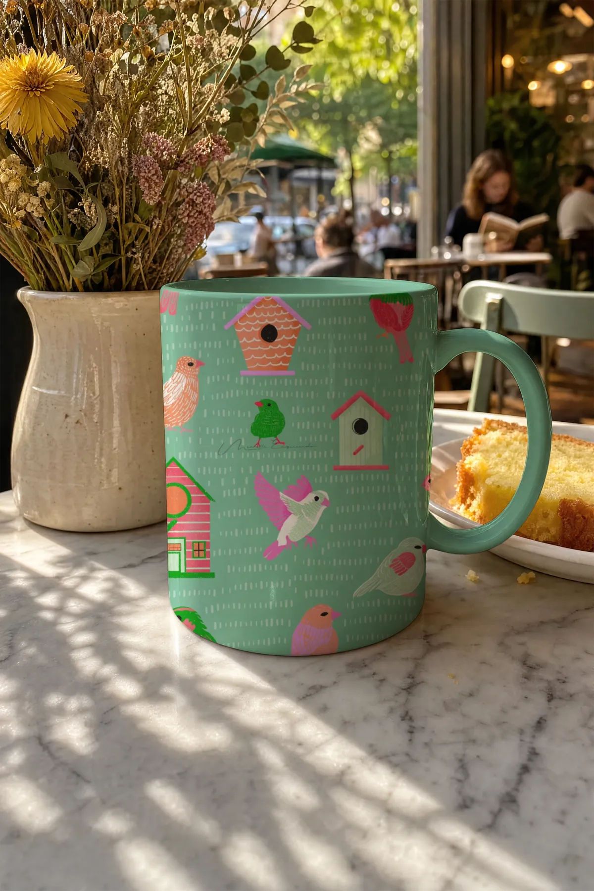 Coffee mug with the Home Tweet Home bird repeat pattern, styled on a marble table in a restaurant with a plate of cake.