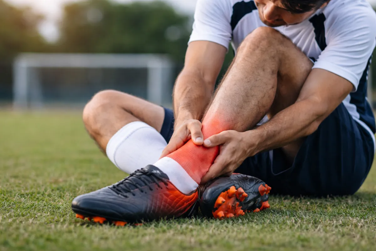 Calciatore seduto sul campo che si tiene la caviglia dolorante dopo una distorsione durante una partita, con evidenza di infiammazione nella zona del malleolo.