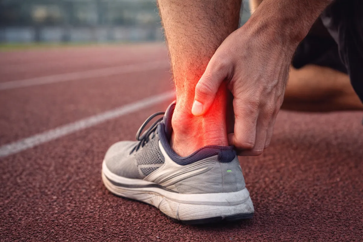 Primo piano di una caviglia su pista da corsa: un runner tiene il tendine d’Achille dolorante, evidenziato in rosso per indicare infiammazione nella parte posteriore del tallone.