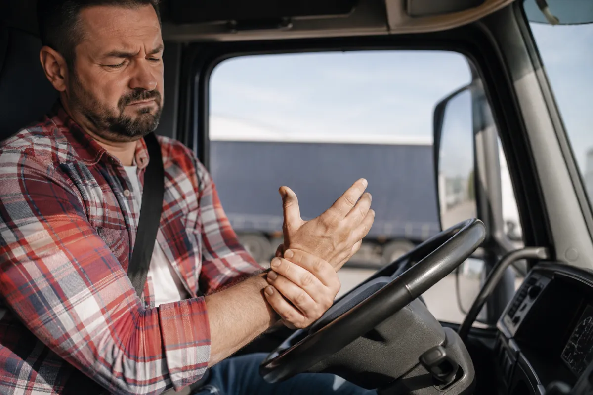 amionista seduto al volante del proprio mezzo mentre si tiene la base del pollice dolorante dopo molte ore di guida, immagine che rappresenta la sesamoidite del pollice, patologia da sovraccarico frequente tra autotrasportatori, tassisti e conducenti professionisti.