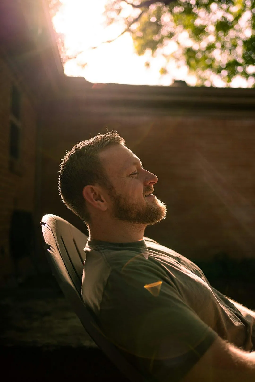 Man seated in profile with eyes closed and soft smile, bathed in dapple sunlight on patio, reflecting peace and intuitive alignment