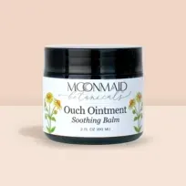 Product photo of MoonMaid Botanicals Ouch Ointment Soothing Balm in a 2 fl oz (60 ml) jar against a soft peach background.