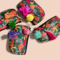 A top-down view of Anna’s travel bags featuring a tropical green and pink floral print. Each zipper bag is decorated with a unique set of colorful accessories, including oversized pink, yellow, purple, and teal tassels and pompoms.