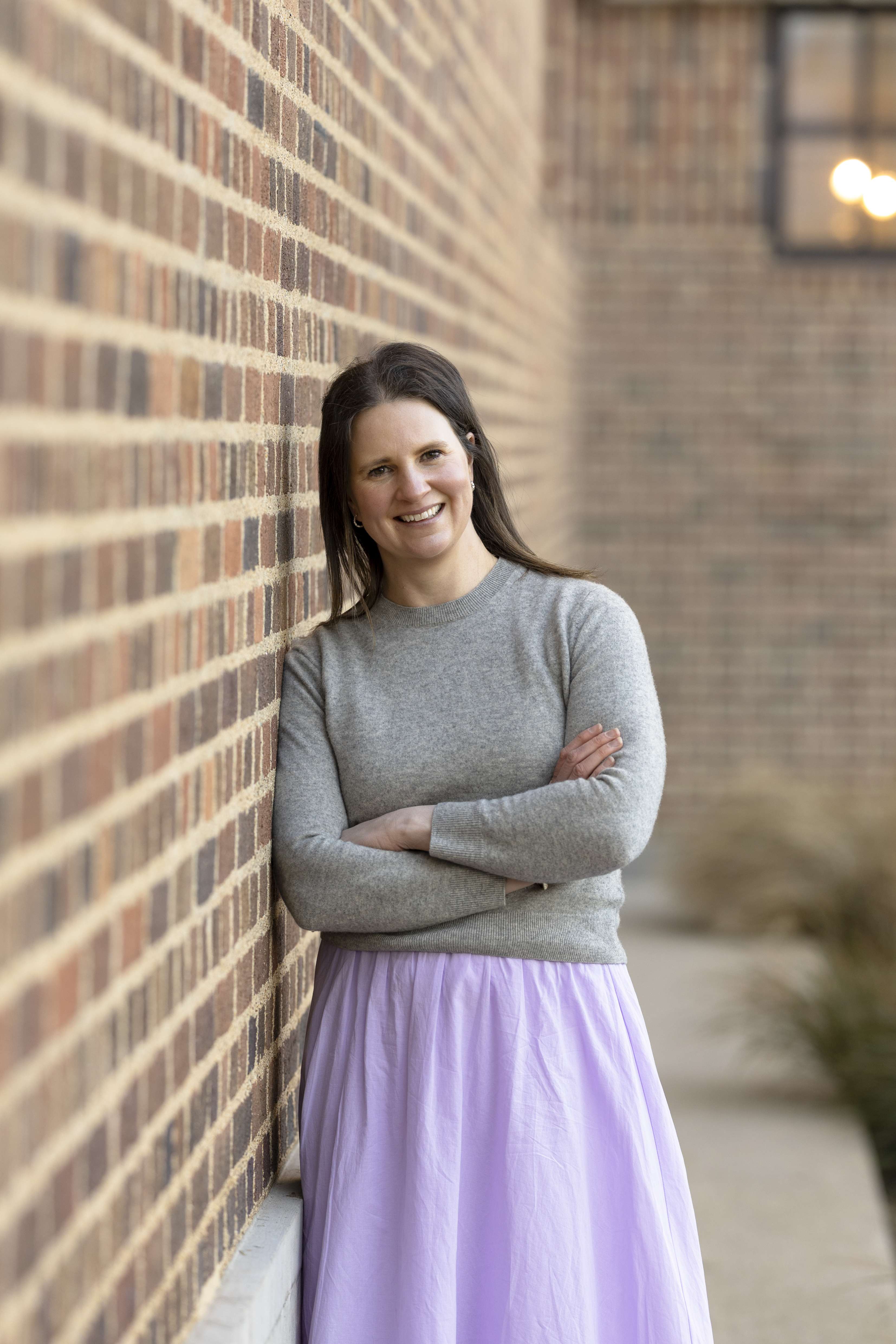 Image of lady leaning against a brown brick wall. She has her head slightly tilted to her right. She has brown hair and is wearing a grey jumper and a lilac skirt.