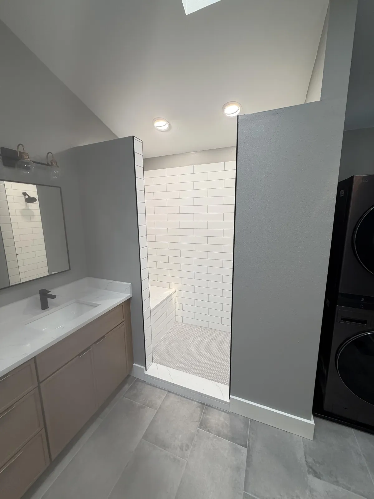 Clean bathroom view showing a light wood vanity with a plant, next to a large glass walk-in shower with black trim.