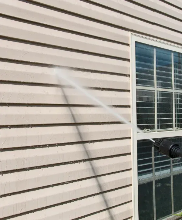 A power washer cleaning light-colored house siding next to a window with blinds inside, seen from an angle.