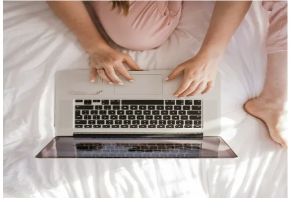 An overhead view of a person in a pink top lying on a white bed typing on a laptop, representing the ease of attending virtual psychiatric appointments from home
