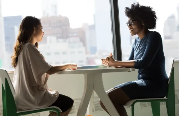 Two women sit across from each other at a white table in a bright office, representing a supportive psychiatric consultation