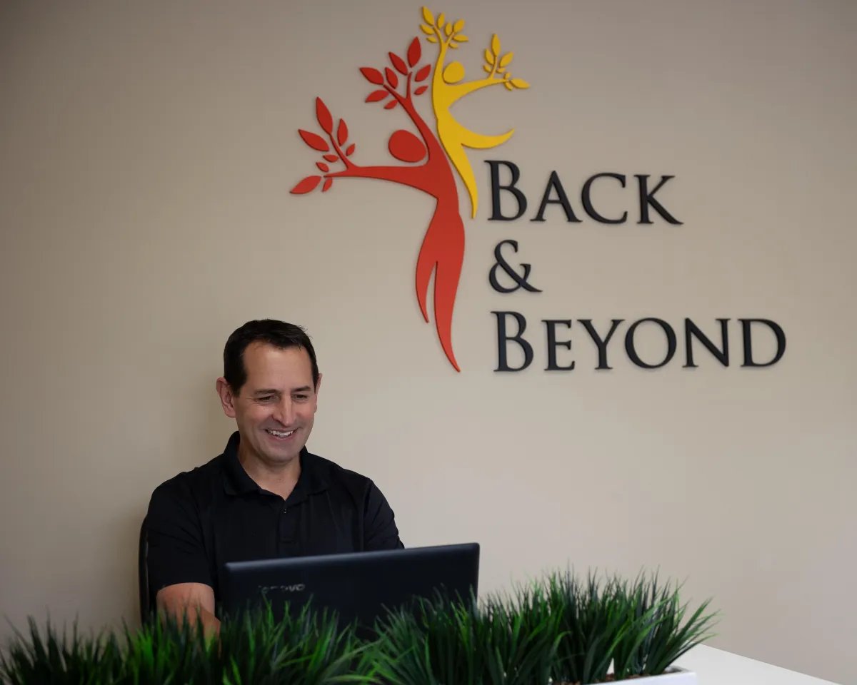 Dr. Craig Burns smiling at reception desk with Back & Beyond logo in background at Redmond chiropractic clinic