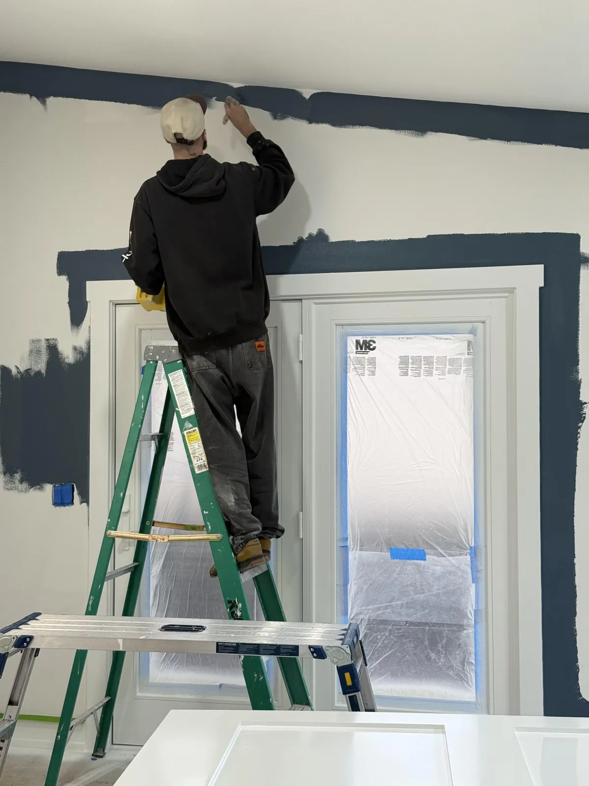 A professional painter in a branded uniform is painting a bright, modern living room wall. Natural light streams in through large windows, highlighting the transformation. The workspace is tidy, with high-quality tools visible, appealing to homeowners seeking a fresh, updated look.