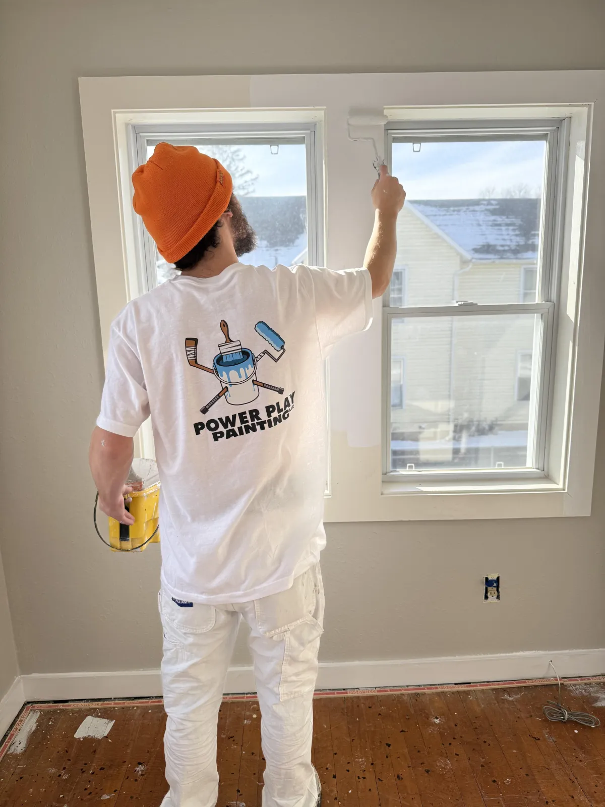 A professional painter in a branded uniform is painting a bright, modern living room wall. Natural light streams in through large windows, highlighting the transformation. The workspace is tidy, with high-quality tools visible, appealing to homeowners seeking a fresh, updated look.