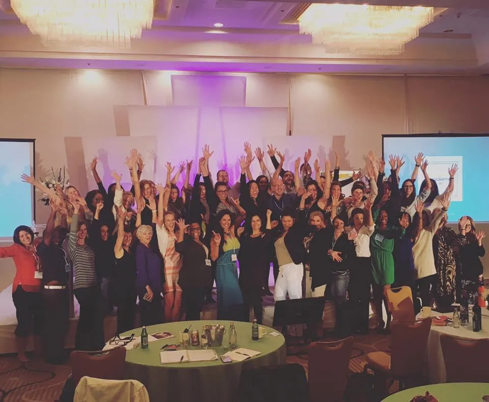 A large group of people standing together in a conference room with their arms raised in celebration. The group consists of men and women of various ages, and they are smiling and looking at the camera. The background includes two large screens displaying presentations and decorative lighting. Round tables with chairs are visible in the foreground, set with notepads, pens, and water bottles.