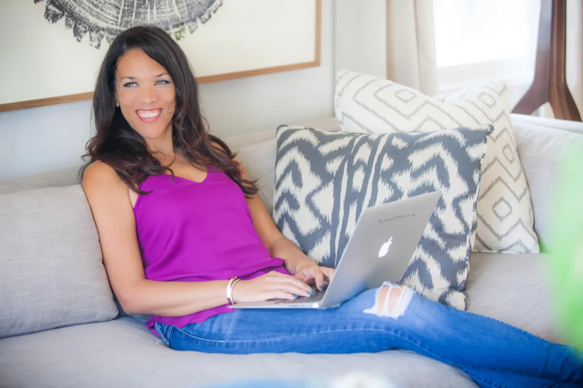 A woman with long dark hair, wearing a bright purple sleeveless top and ripped blue jeans, is reclining on a couch with a laptop on her lap. She is smiling and looking at the camera. The couch has patterned cushions, and there is a framed artwork on the wall behind her.