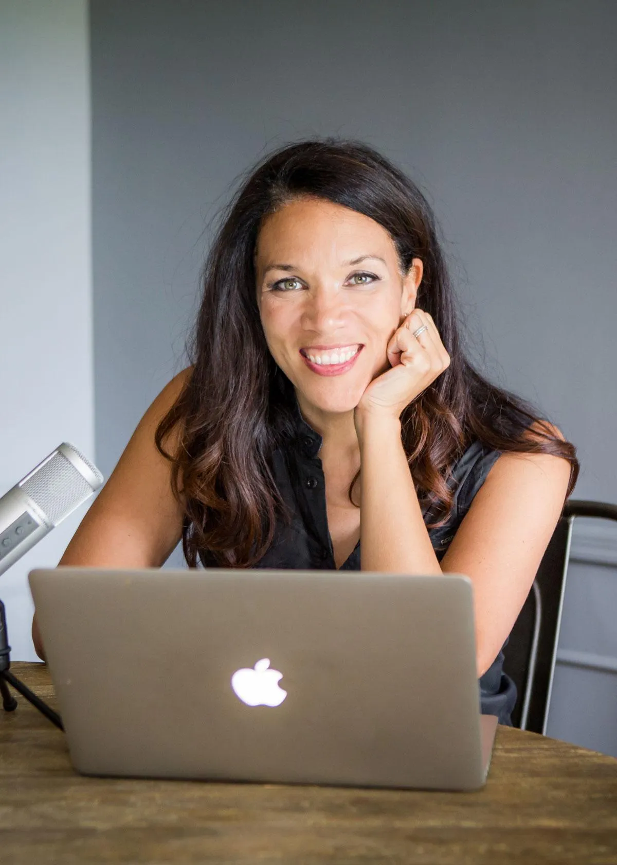 A woman with long dark hair, wearing a sleeveless black top, sits at a wooden table with a laptop in front of her. She is smiling and resting her chin on her hand. A microphone is positioned on the table to her left.