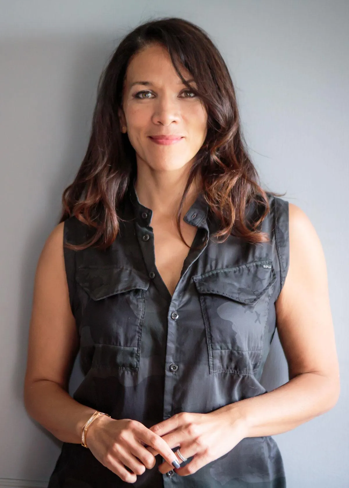 A woman with shoulder-length dark hair, wearing a sleeveless black shirt with buttoned pockets, standing in front of a plain gray background. She is smiling gently and has her hands clasped in front of her.