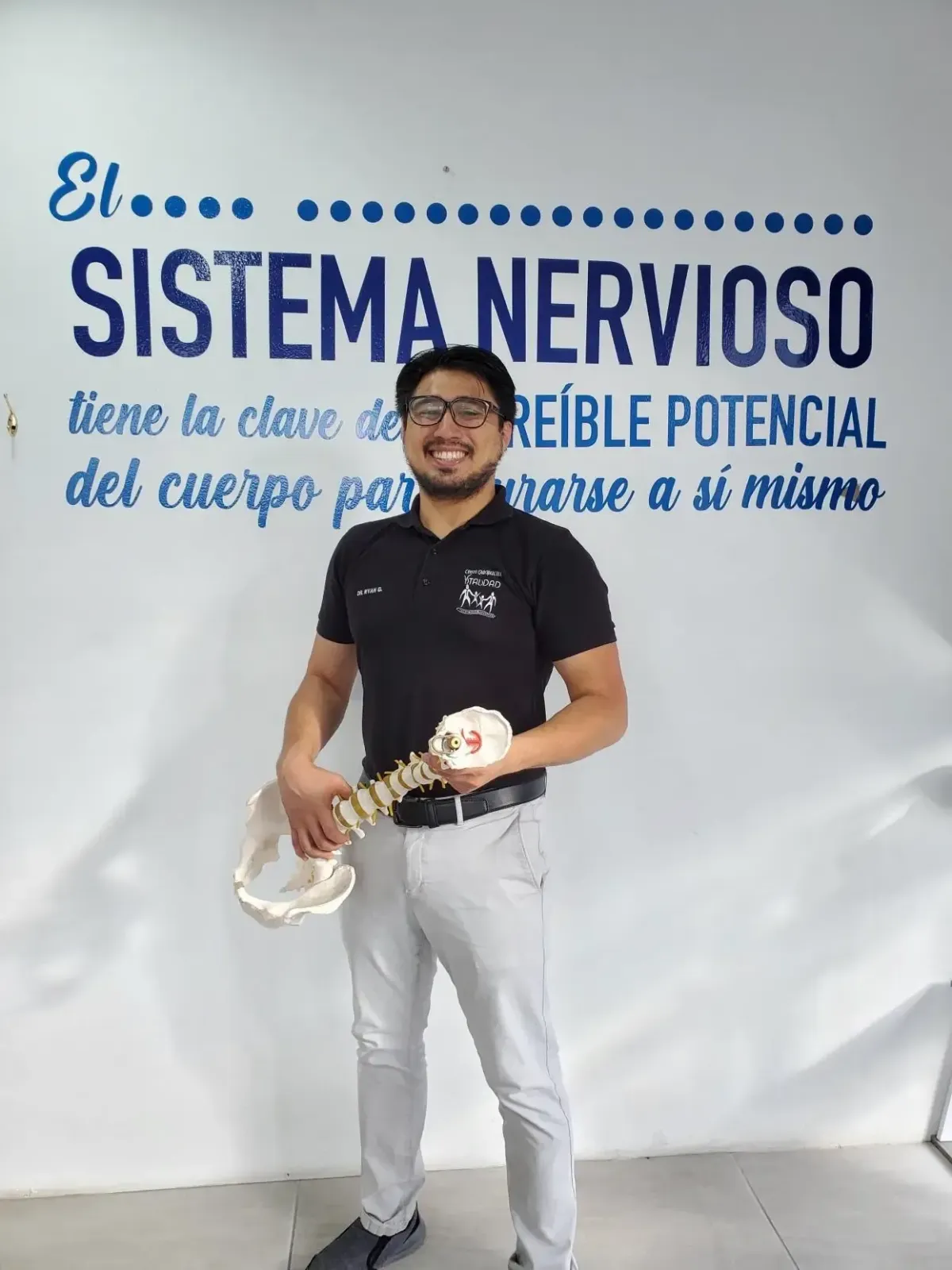 Dr. Ryan García sonriente sosteniendo una columna vertebral con fondo informativo sobre el sistema nervioso, educando sobre la importancia de la quiropráctica para la salud del cuerpo
