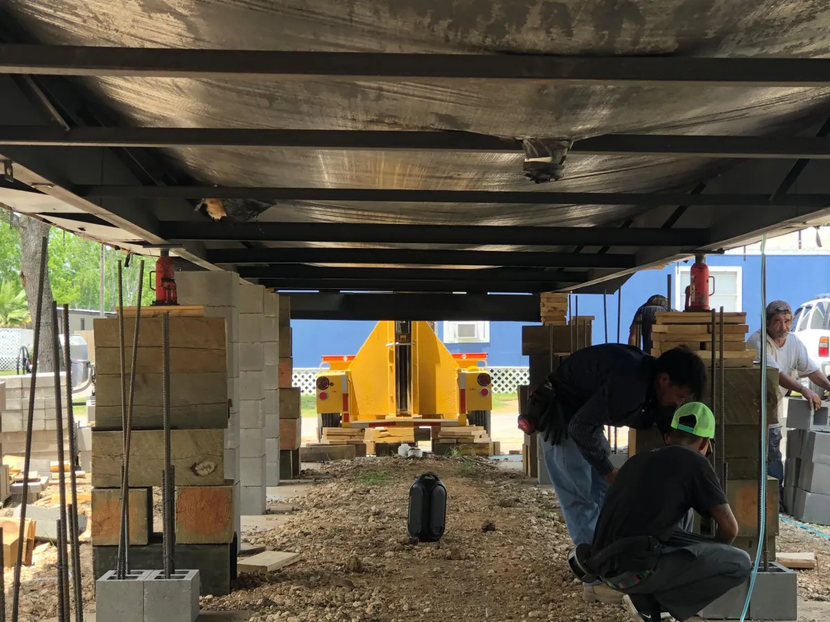 Workers performing mobile home leveling, positioned underneath the structure supported by stacked concrete blocks and wooden beams, with hydraulic jacks and rebar reinforcements visible in the process. A yellow hydraulic machine is in the background.