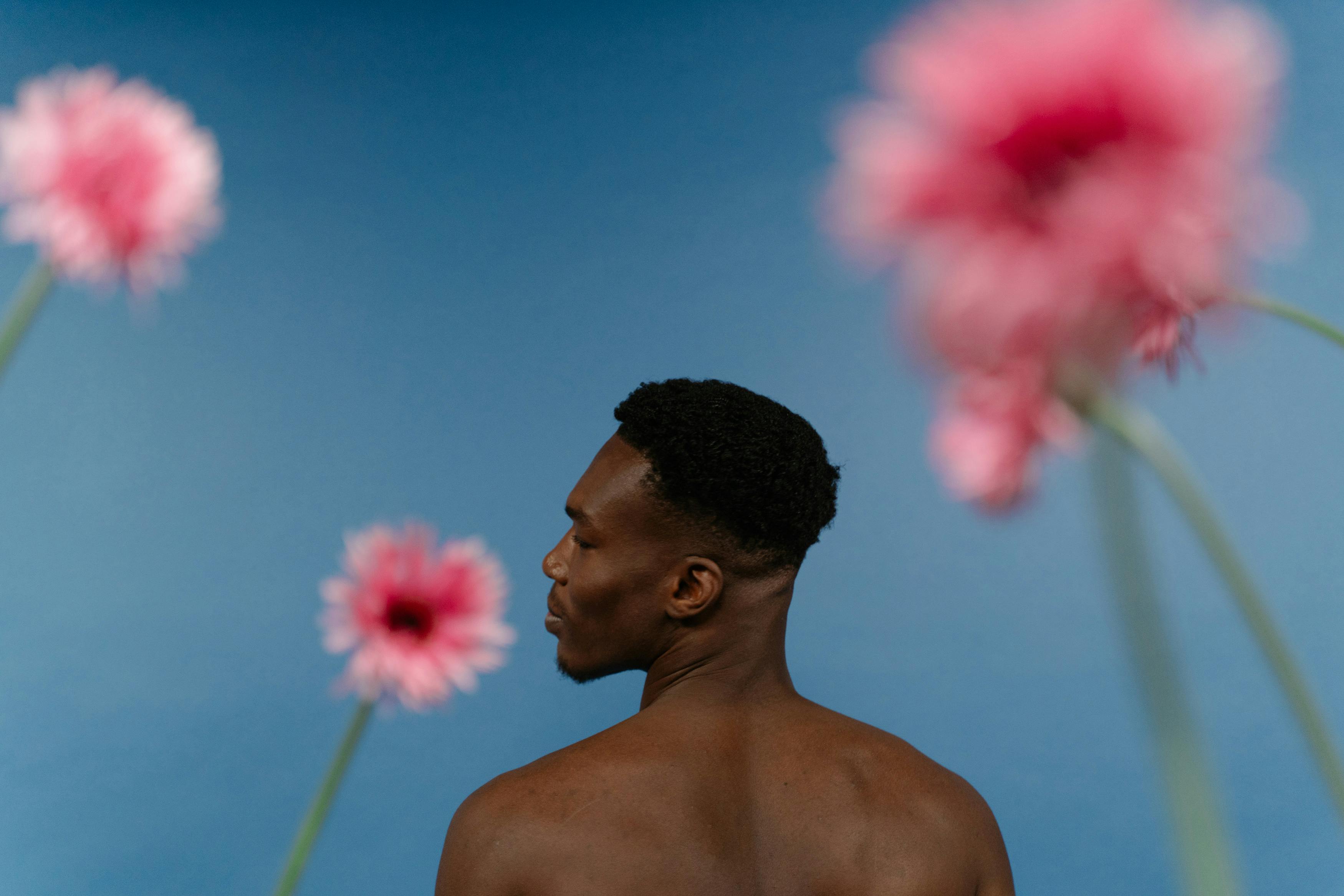 A shirtless Black man photographed from the back and side profile against a soft blue background with large blurred pink flowers in the foreground and background. The image has an artistic and minimal aesthetic, with soft lighting and shallow depth of field creating a calm, editorial-style atmosphere.