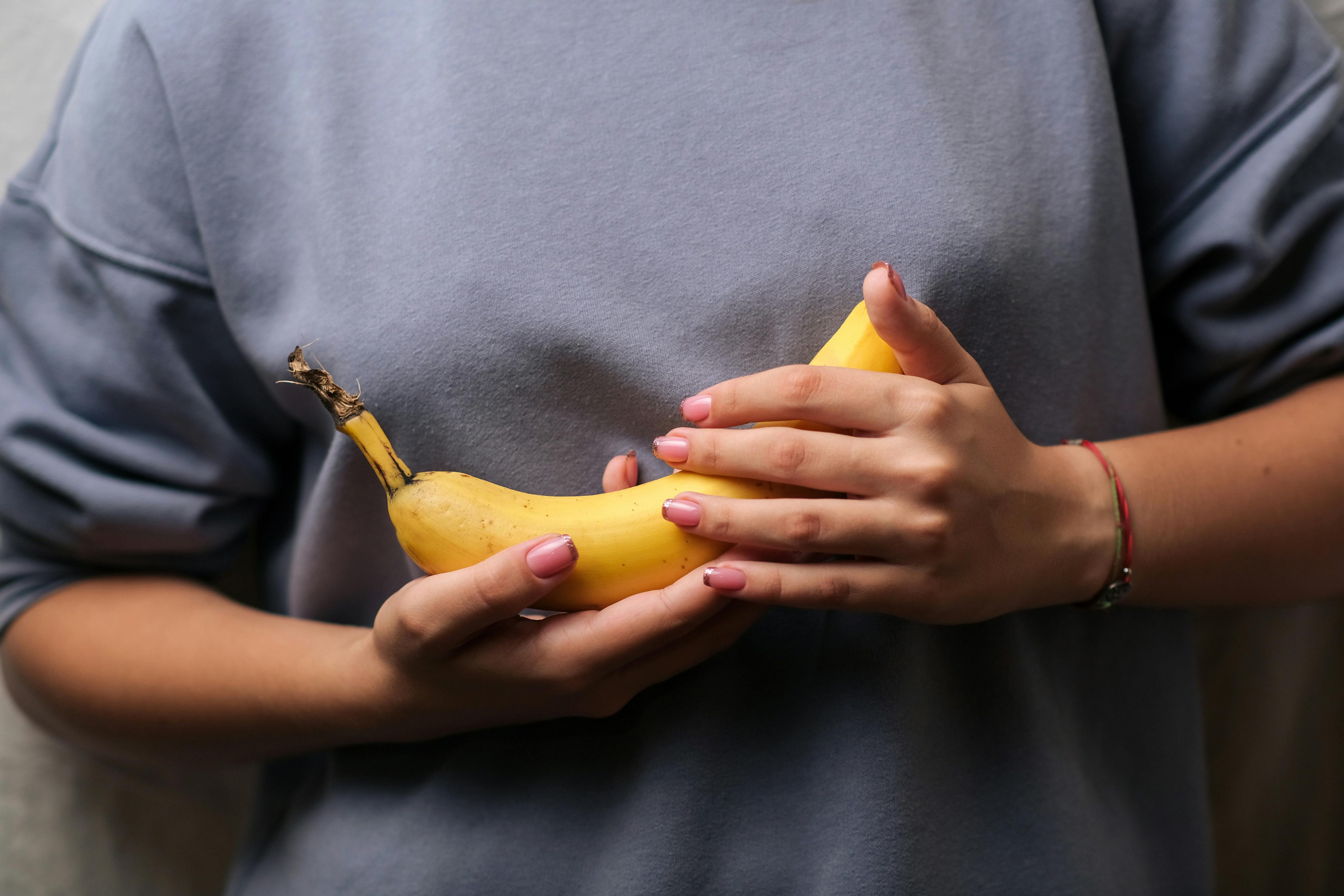 A person wearing a loose blue sweatshirt holds a ripe yellow banana with both hands at chest level. The image is cropped to show only the torso and hands, with the banana positioned horizontally in the center of the frame. The person has light pink nail polish and a thin bracelet on one wrist. The background is softly blurred, drawing attention to the banana and the hands holding it.