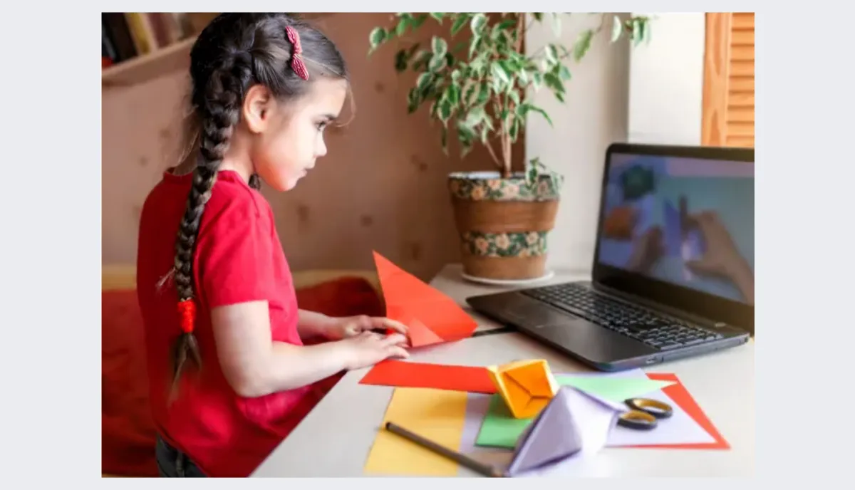 Girl folds paper while watching teacher on laptop