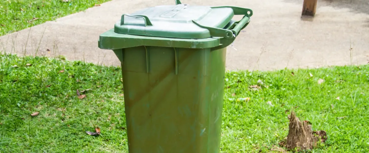 Green outdoor trash bin placed on grass near concrete walkway