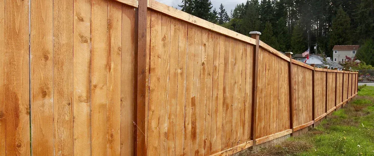 A long, newly built wooden fence stretches across a grassy area with houses and trees visible in the background.