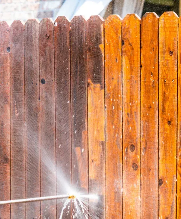 A close-up of a wooden fence being pressure washed, showing a clean, bright section next to a dirty, dark one.