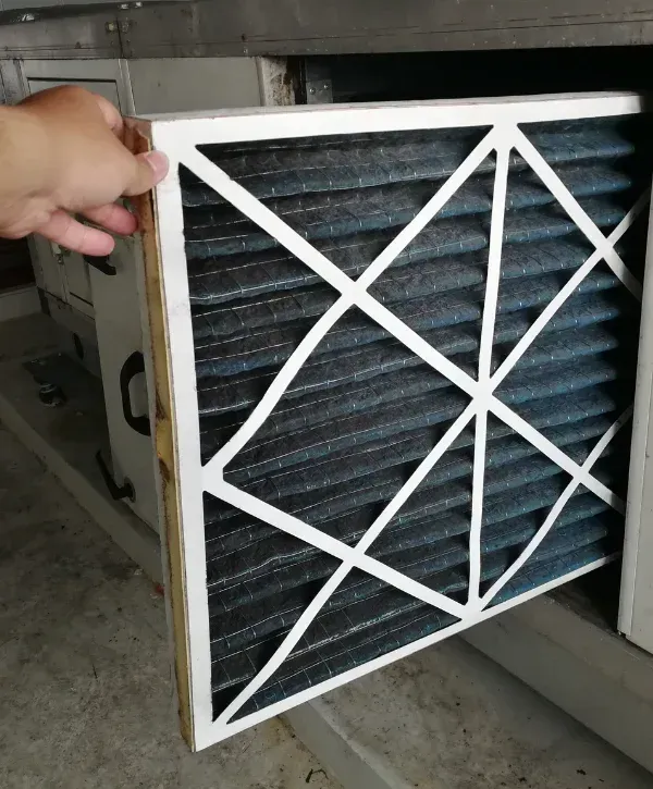 A hand pulls out a dirty, pleated blue air filter from an HVAC unit, revealing debris on its surface.