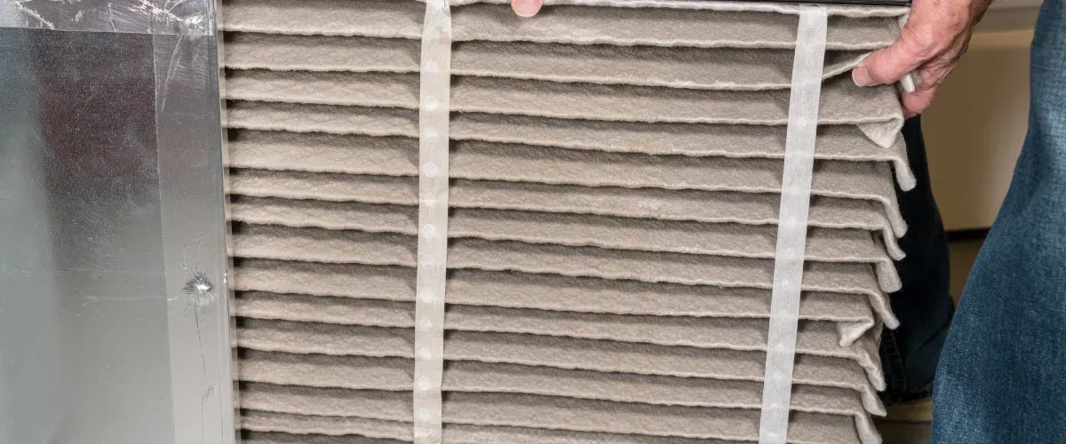 A person's hands installing or removing a dirty pleated air filter from an HVAC unit, with silver ductwork visible.