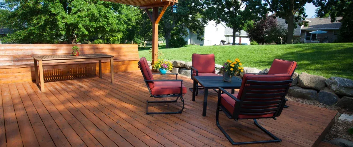 A spacious wooden backyard deck with a table, built-in bench, and four chairs with red cushions, surrounded by green trees and grass.