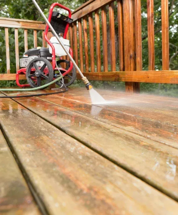 A close-up shot of an old wooden deck being cleaned by a pressure washer, with a red pressure washer machine in the background.