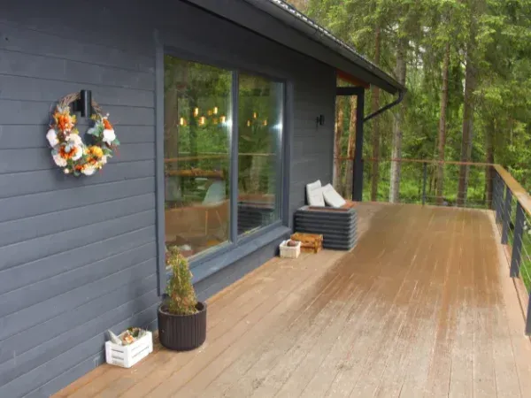 A wooden deck extends from a modern grey house into a lush forest, featuring a wreath on the wall and seating.