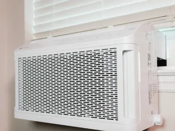A close-up, angled view of a white window air conditioner with a grated front panel, installed in a window with blinds.