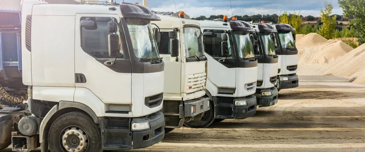 A line of white semitrucks parked on a dirt lot, with large piles of sand in the background.